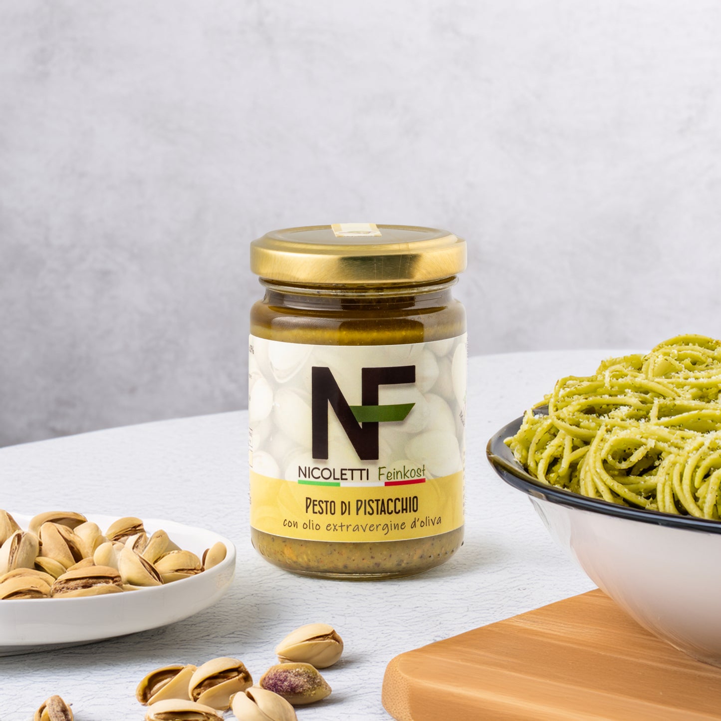 Ein Glas Pesto di Pistacchio aus Sizilien, exklusiv für Nicoletti Feinkost hergestellt, steht neben losem Pistazien und einigen in einem Teller. Rechts ist ein Teller Spaghetti, verfeinert mit dem Pesto und bestreut mit Parmesan – ein authentisches sizilianisches Genusserlebnis.