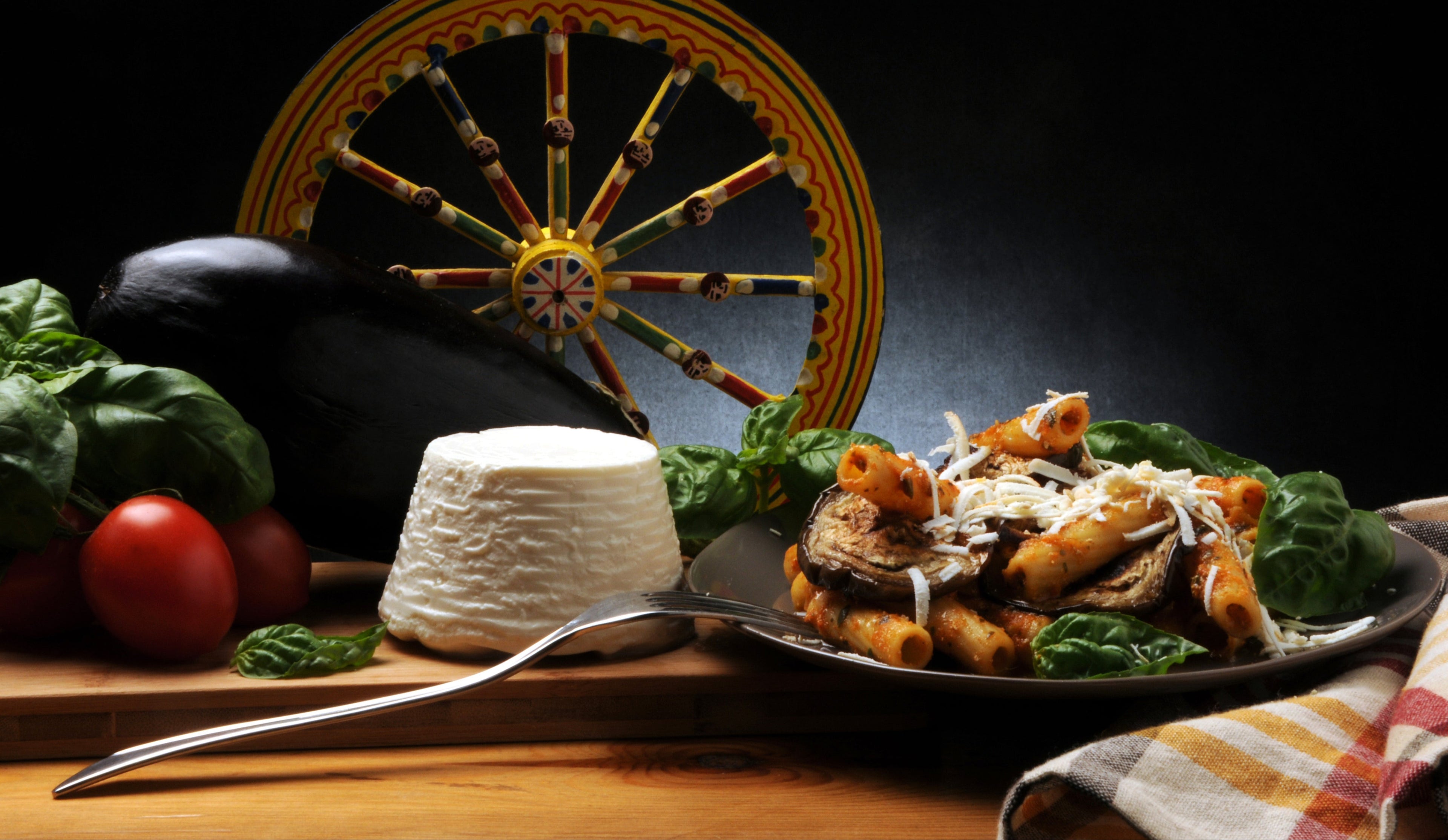 Sizilianische Spezialitäten: Ricotta Salata, Rigatoni alla Norma, frische Tomaten, Auberginen und ein traditionelles Carretto Siciliano, die das italienische Flair vermitteln